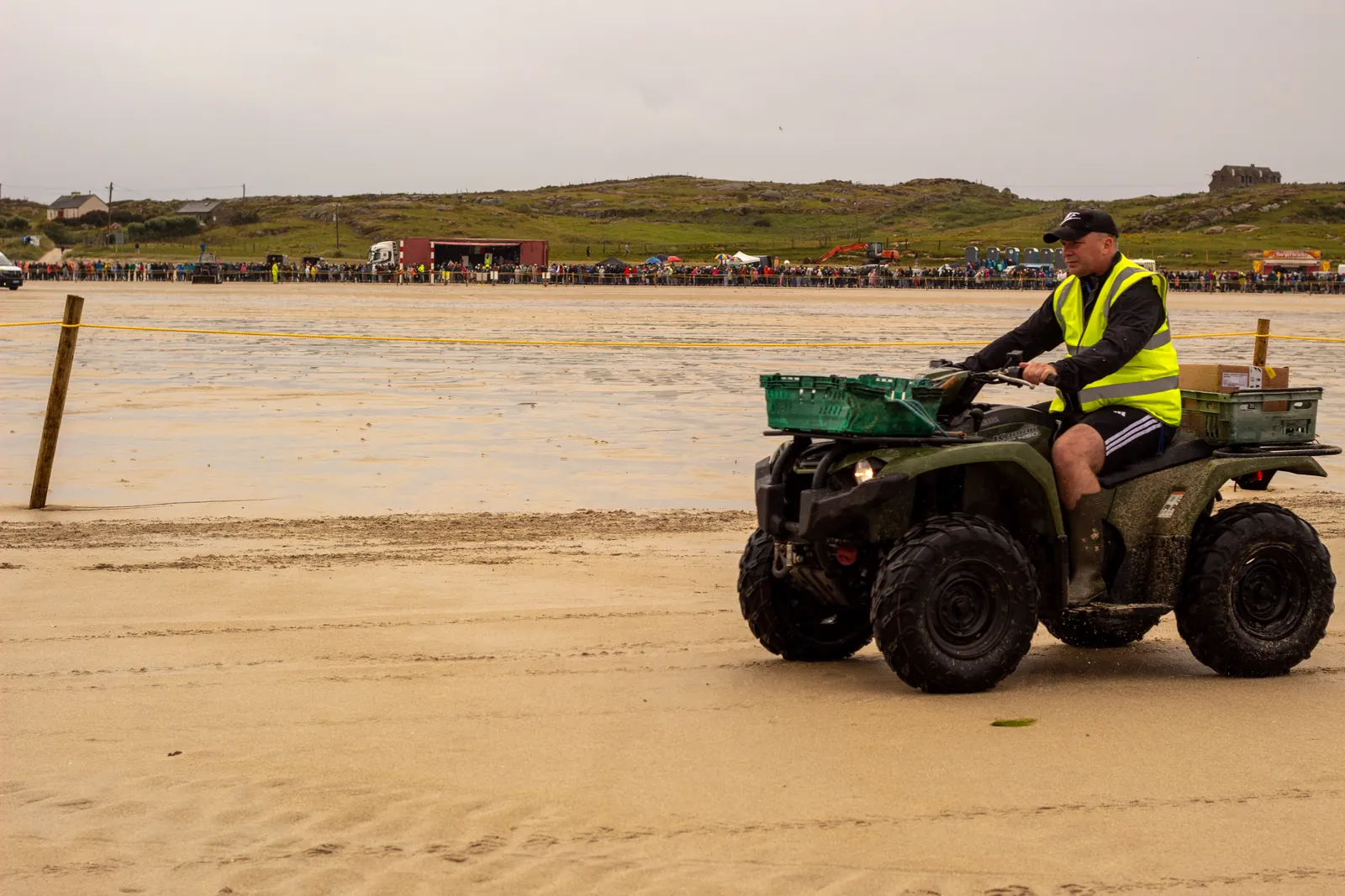 A steward in a high-visibility vest rides a quad bike loaded with crates along the sand beside the roped-off course.