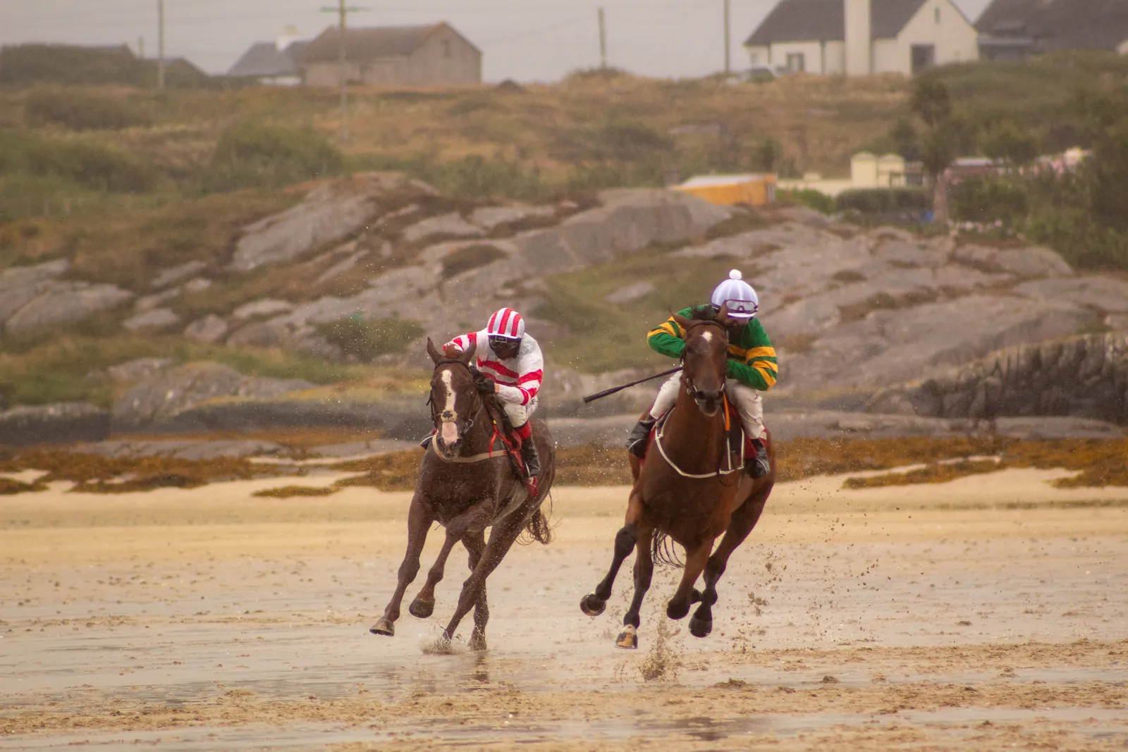 Two horses race neck and neck towards the camera in the rain, one jockey in green and gold, the other in red and white stripes, granite outcrops and cottages behind them.