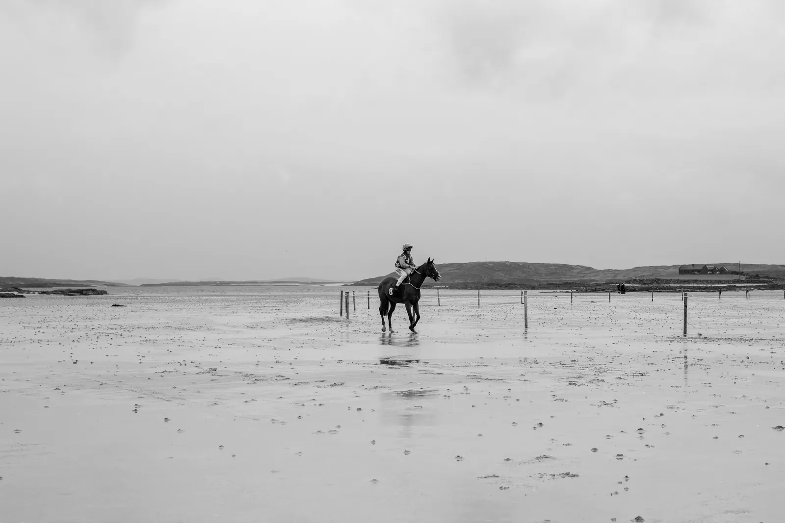A single rider and horse stand alone in the middle of the enormous flat strand, reflected in a sheet of shallow water.