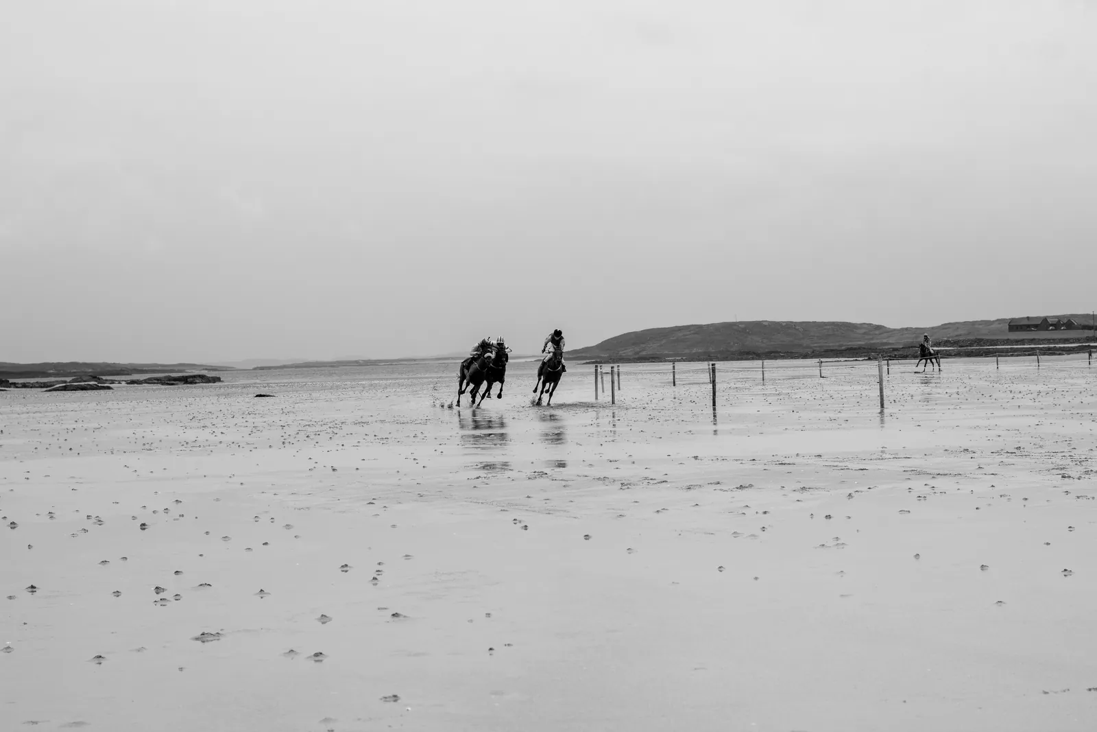 Two horses race side by side, tiny in the frame, along the far edge of a vast expanse of wet sand at Omey with the sea behind them.