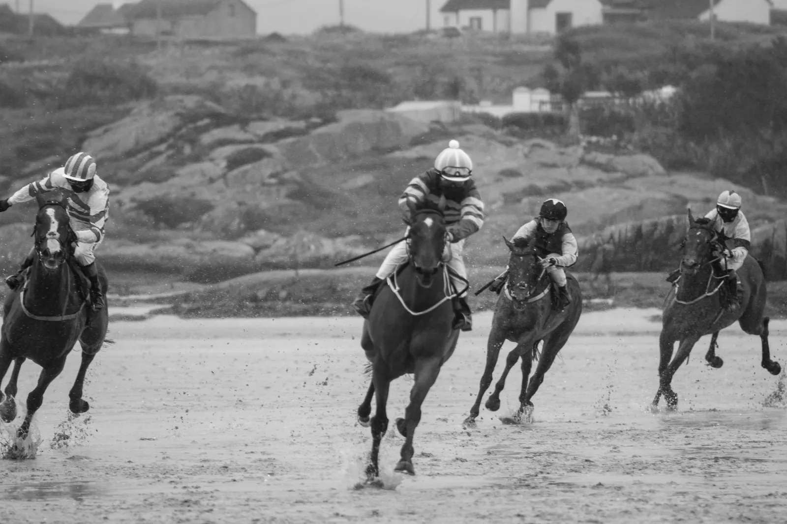 Five horses spread across the strand galloping towards the camera through falling rain, riders low over their necks, spray coming off the sand.