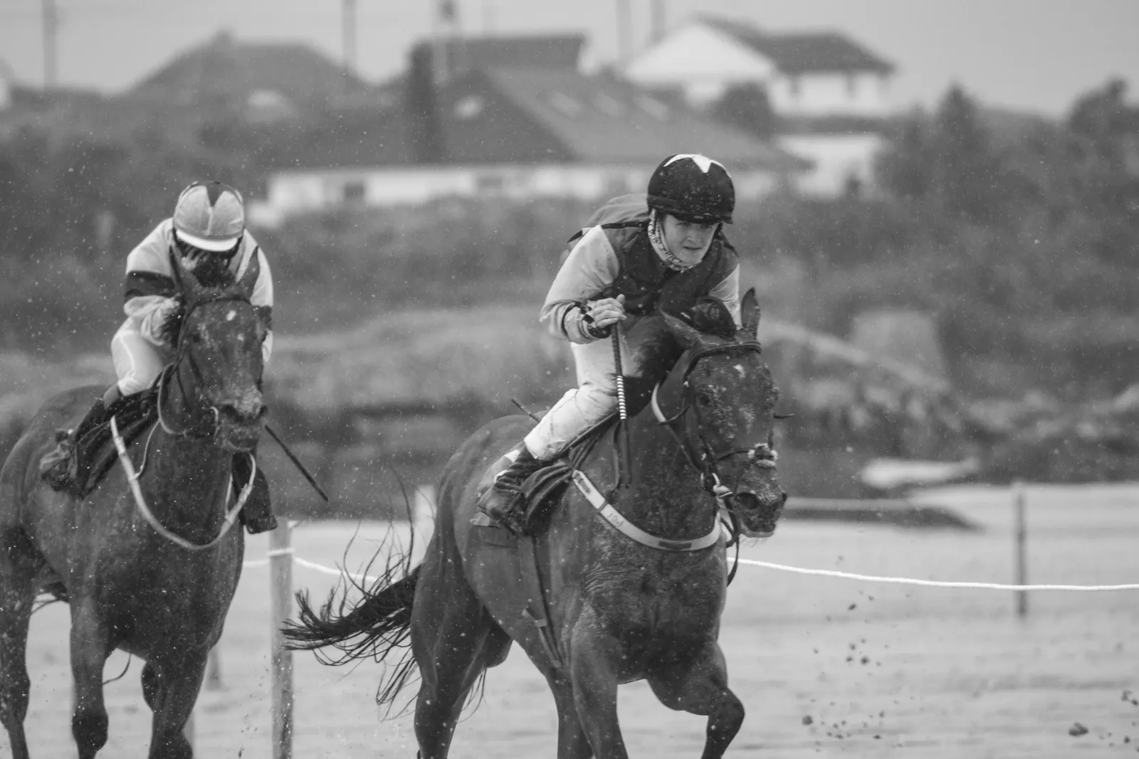 Close-up of a rider driving a dark horse forward through heavy rain at Omey, a second horse and rider just behind, raindrops visible across the frame.